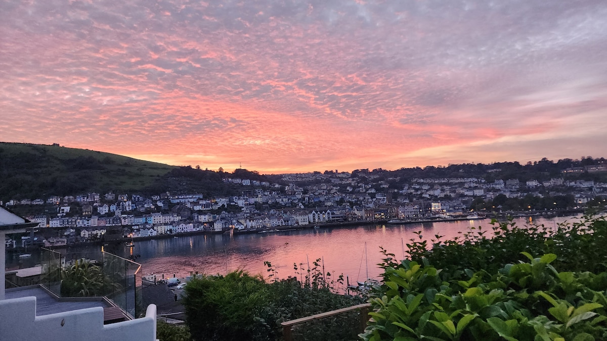 A stunning sunset casts vibrant hues across the sky, reflecting on the calm waters of the River Dart. The treed foreground includes lush bushes, while the charming village beyond features rows of buildings and the scenic green hills in the distance.