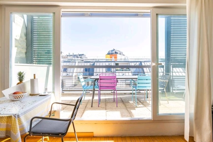 Large glass doors open to a balcony featuring colorful outdoor furniture, including chairs and a dining table. The bright area is illuminated by natural light, with a view of buildings in the distance and a soft blue sky overhead.