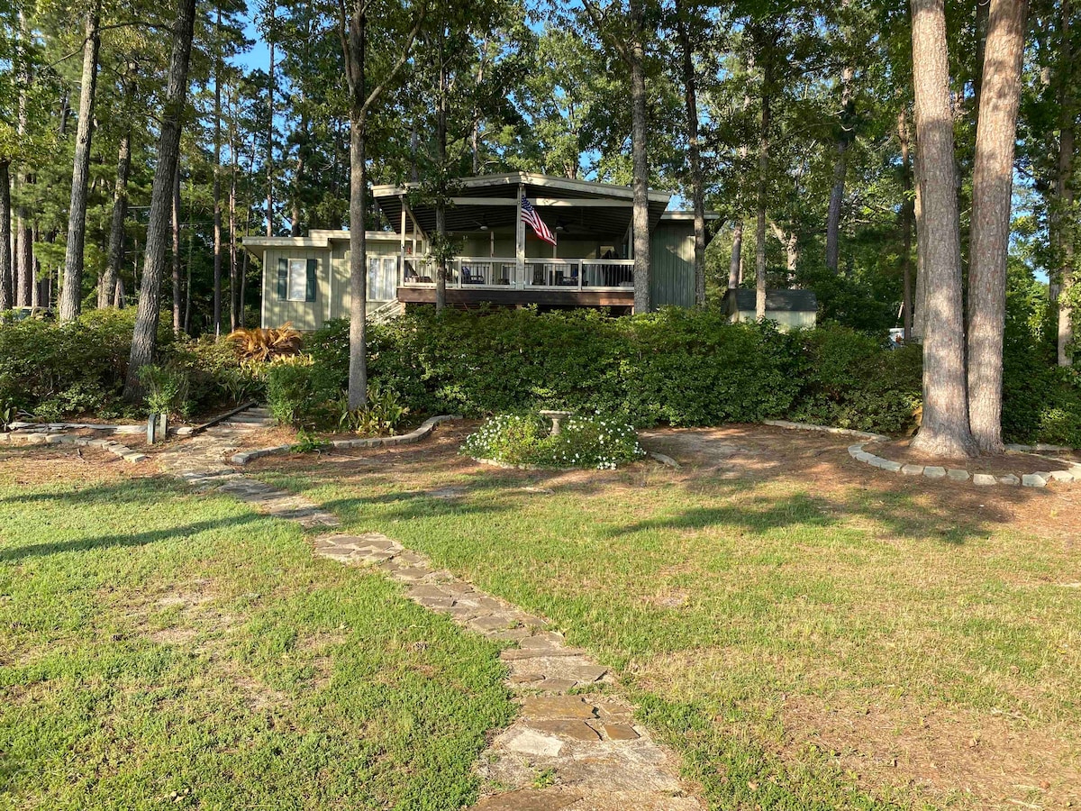 A tranquil lakeside home is surrounded by trees, featuring a covered porch with an American flag. A stone pathway leads from the well-kept lawn to the entrance, highlighting the natural landscape and inviting atmosphere.