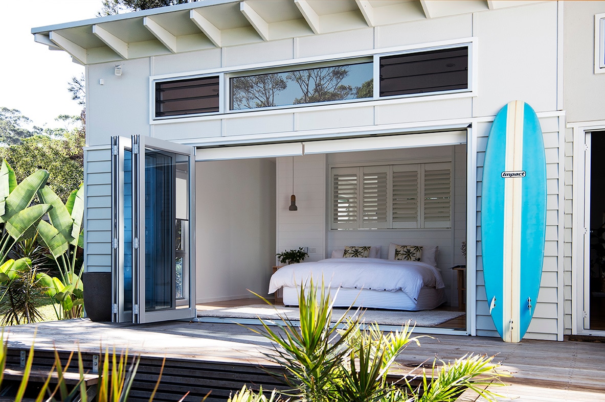 An open bedroom is visible through large bi-fold doors, featuring a spacious bed dressed in neutral linens. A surfboard leans against the house, complemented by green tropical plants in the foreground. Natural light fills the room, creating a relaxed coastal ambiance.