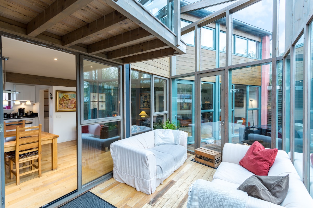 A light-filled living area is enclosed by expansive glass walls, featuring comfortable sofas and a wooden dining table. The open concept connects to the kitchen, while a wooden floor enhances the natural ambiance. Soft furnishings add subtle texture to the inviting space.