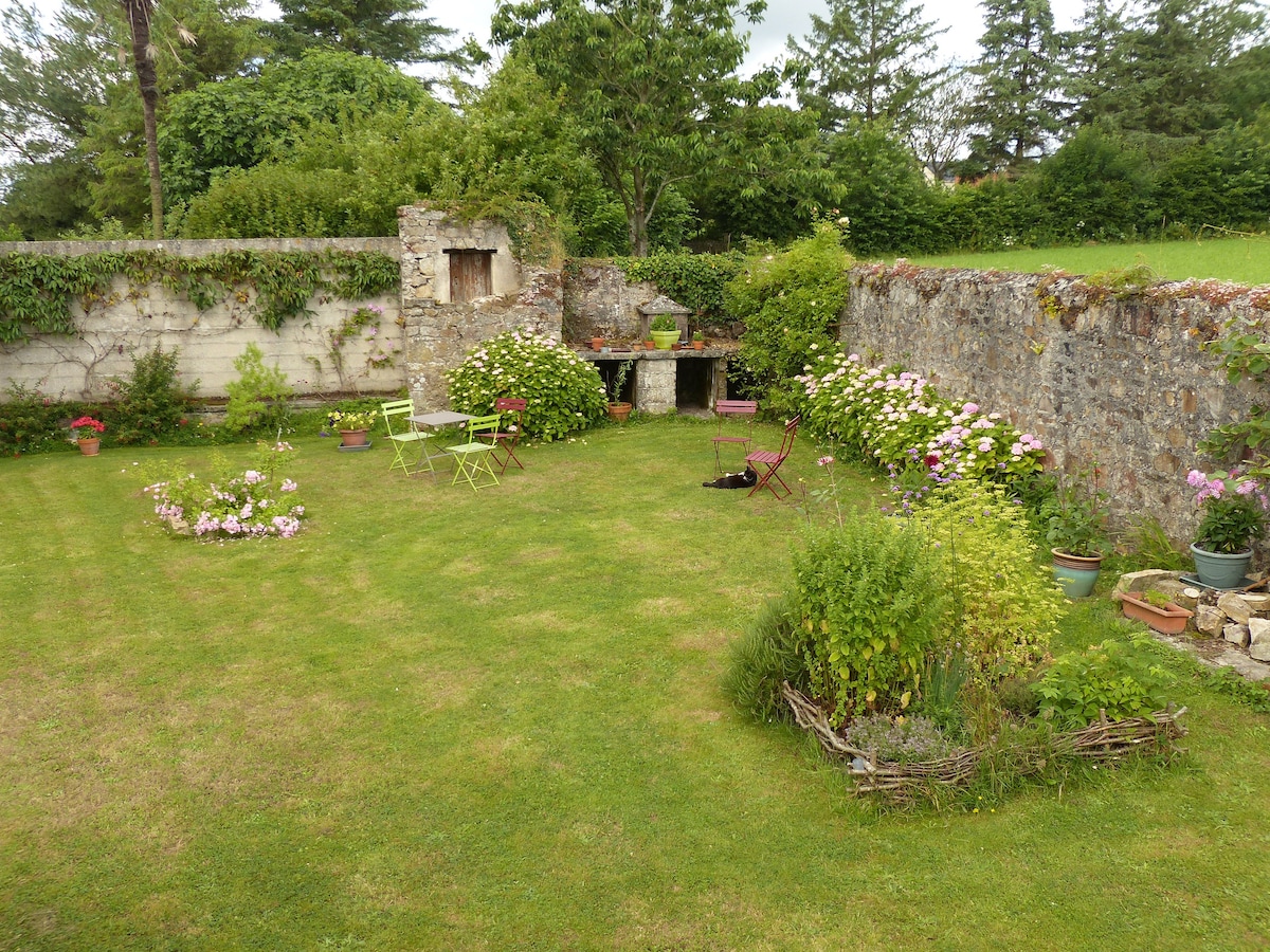 A private garden is surrounded by low stone walls, featuring well-tended flower beds with various plants in pots. A small stone structure with an opening is visible. Green chairs are placed around a table, creating a quiet space for relaxation amidst the greenery.