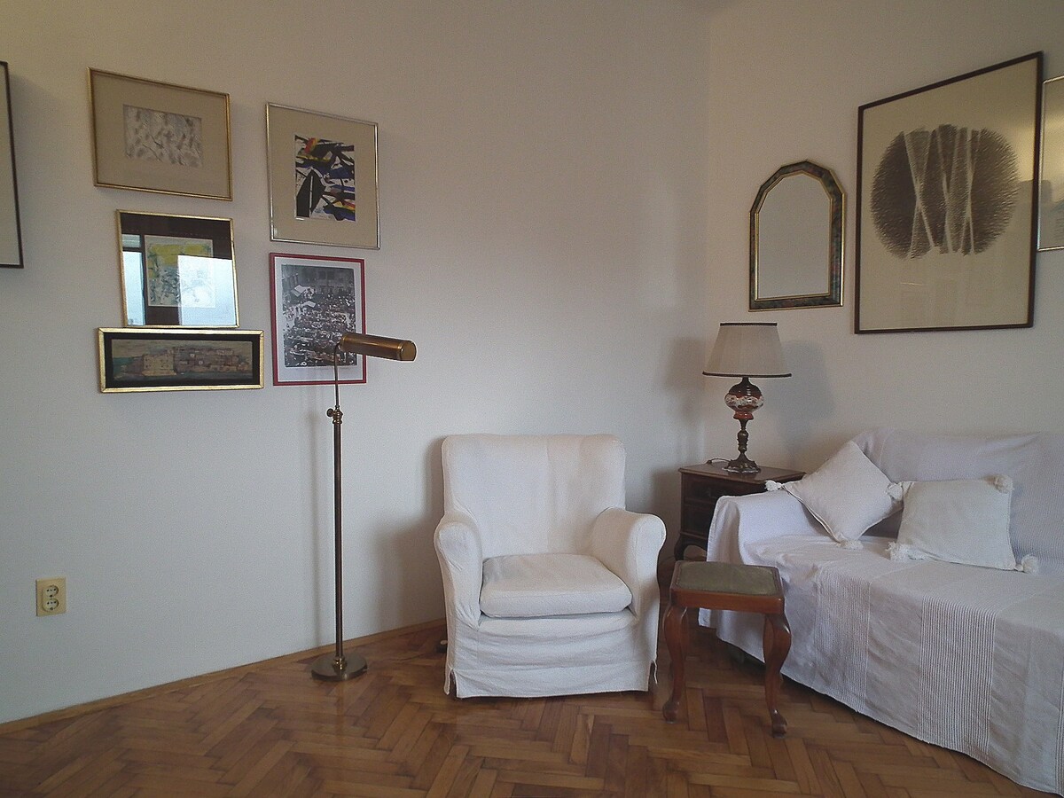 A living area is presented, featuring a white armchair and a sofa draped in a white cover. Decorative framed artwork adorns the walls, and a small wooden side table holds a lamp with a white shade. Warm wood flooring complements the space.