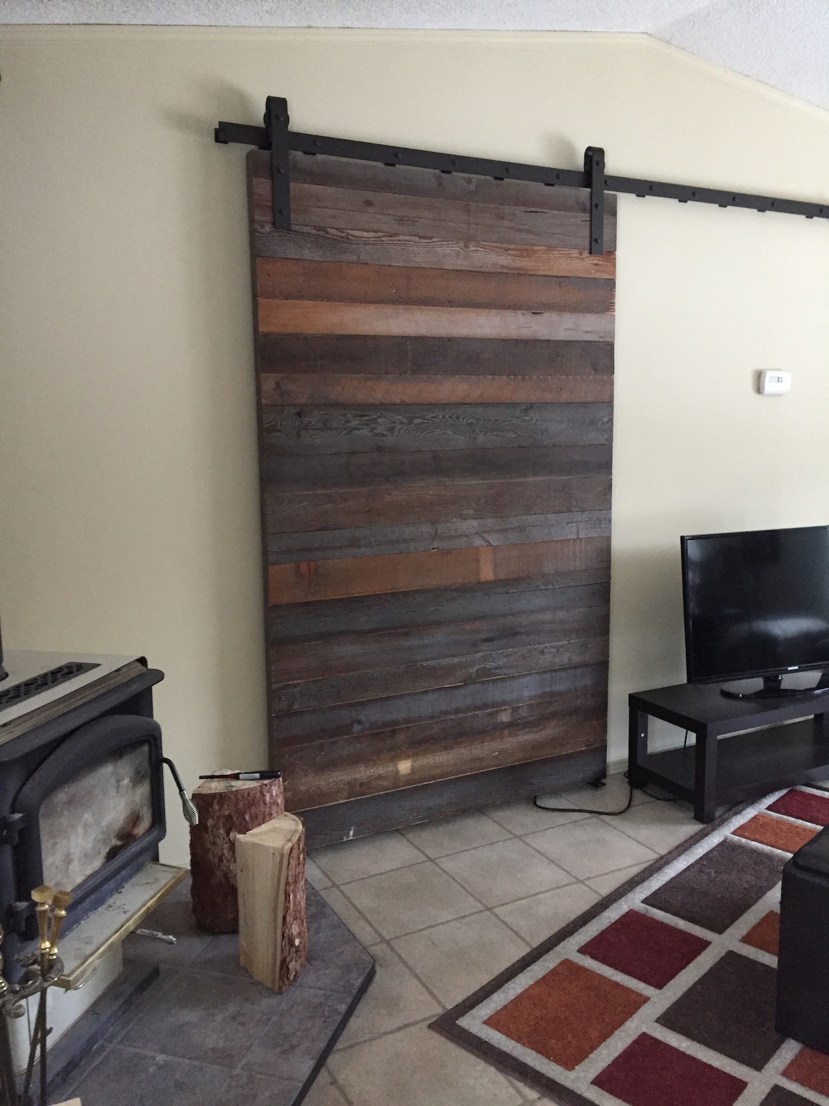 A rustic barn door, crafted from varying shades of wood, is mounted on a black metal track. The door stands beside a cozy wood stove, with a small log pile beside it. A contemporary black television rests on a simple table nearby, set against a neutral wall.