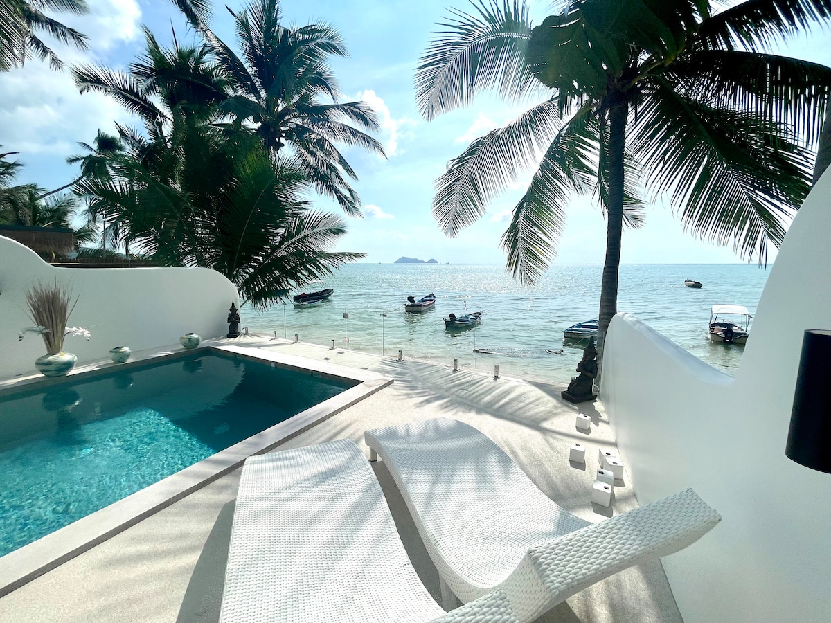 A serene outdoor space features a swimming pool surrounded by lounge chairs, facing the beach. Palm trees provide shade as boats rest on the calm water, and the gentle waves reflect sunlight. The scene offers a direct view of the shoreline, enhancing the tranquil ambiance.