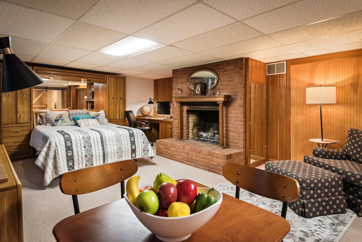 A cozy suite is presented with wood paneling and a comfortable sleeping area featuring a patterned quilt on the bed. A dining table with a bowl of colorful fruit is positioned in front, while a fireplace adds warmth to the room. Seating is provided by an upholstered chair.