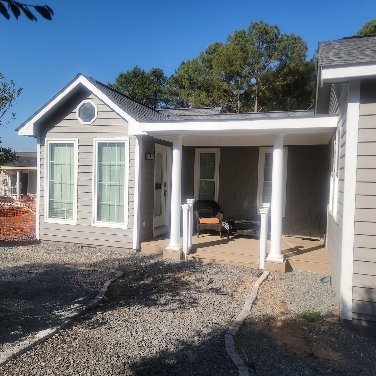 The exterior of the apartment features a light gray facade with white trim and large windows. A covered porch is supported by classic columns, and a cozy seating area is visible on the porch. Surrounding greenery adds a touch of nature to the setting.