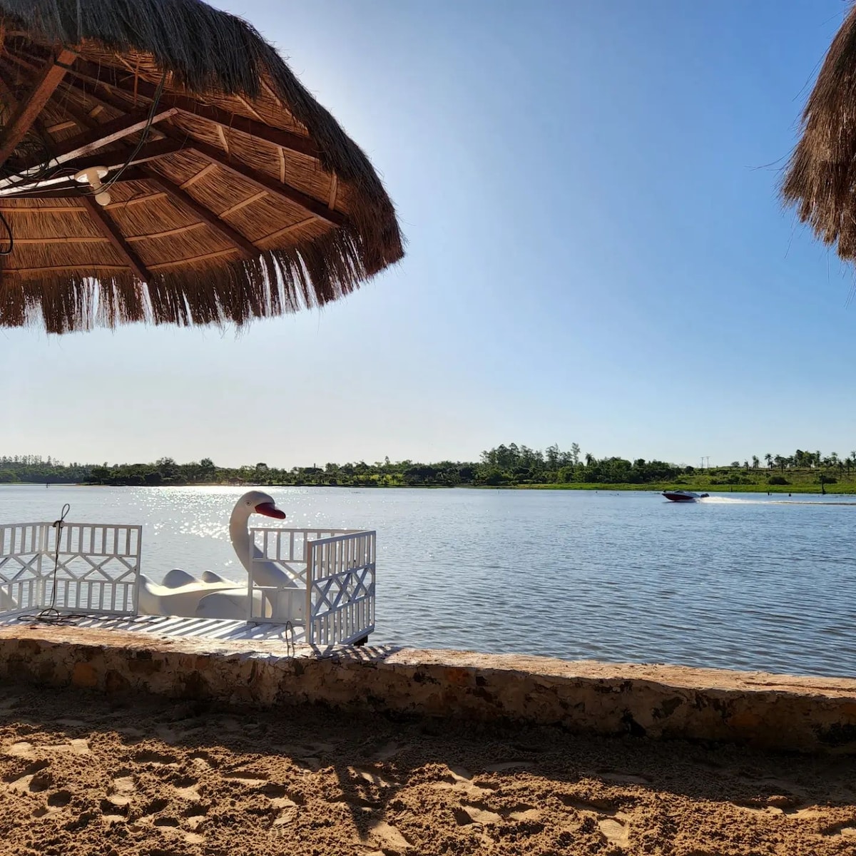A serene view of a calm lake is framed by a thatched umbrella, providing shade. A swan-shaped float sits at the water's edge, while lush greenery lines the opposite shore. A boat glides across the lake in the background, under a clear blue sky.