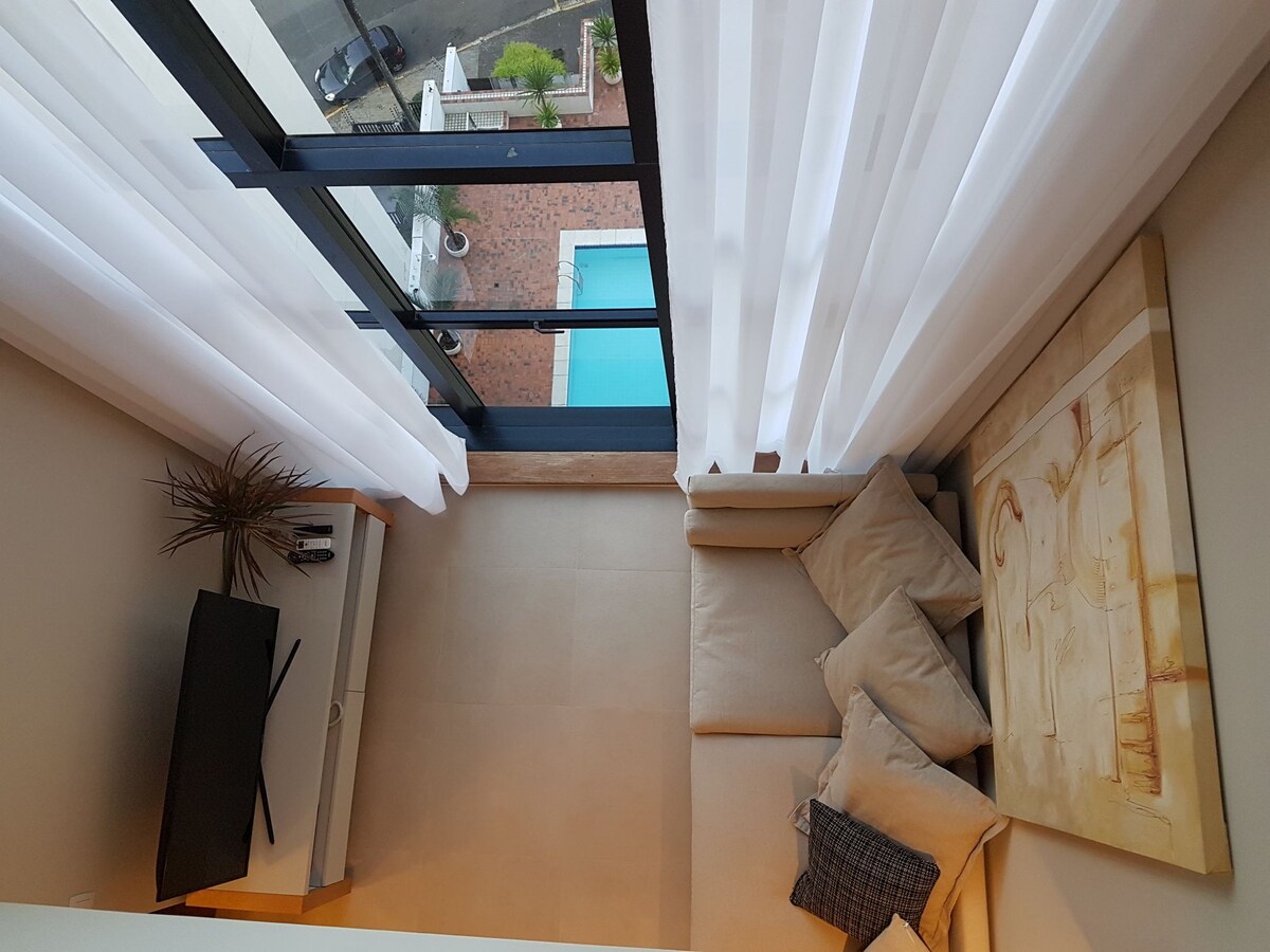 An aerial view of a cozy living area features a sofa adorned with various cushions, positioned next to a modern TV. Sheer white curtains allow natural light to diffuse softly, framing a glimpse of a pool area outside through large glass doors.