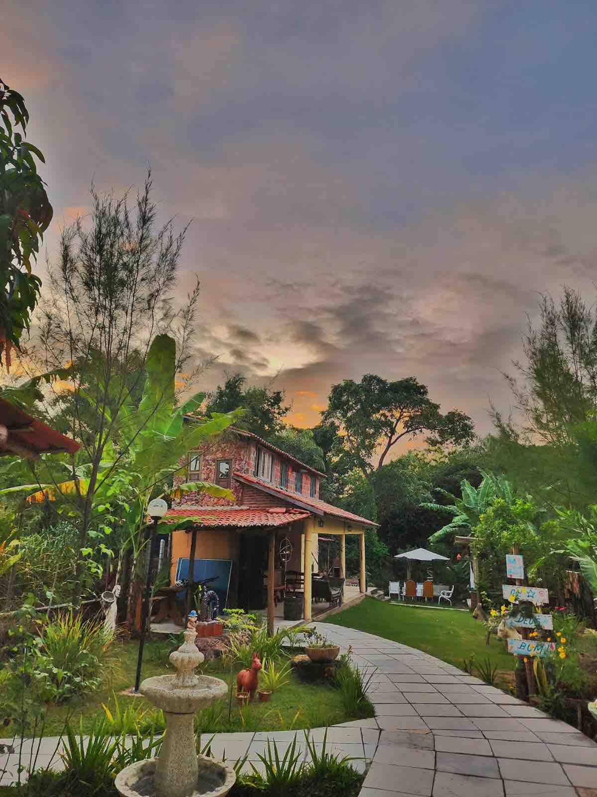 A charming pathway meanders through lush greenery, leading towards a two-story structure with a red-tiled roof. The sky is adorned with soft hues of orange and blue, and a decorative fountain is visible in the foreground, surrounded by vibrant plants.
