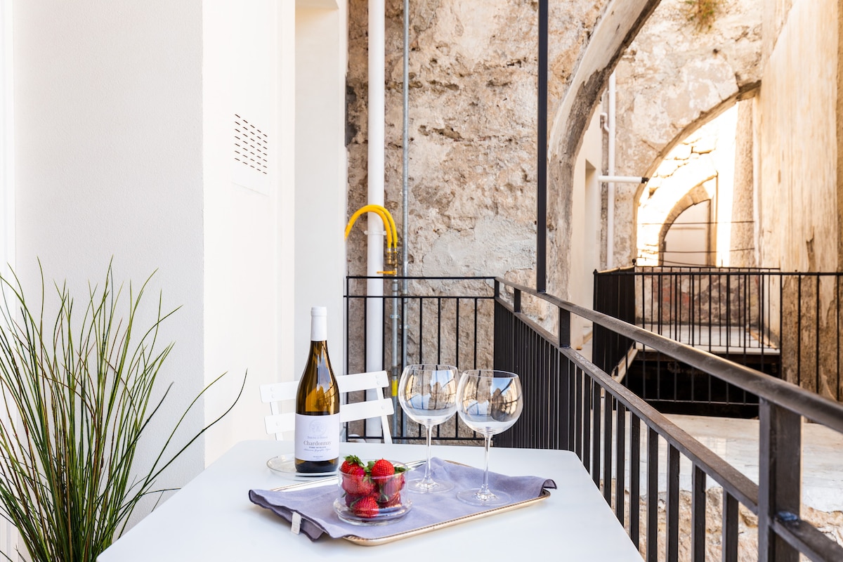 A small outdoor dining area is presented, featuring a table set with a bottle of wine and elegant glasses. Fresh strawberries are arranged on a tray. The backdrop showcases stone walls, adding texture and character to the space.