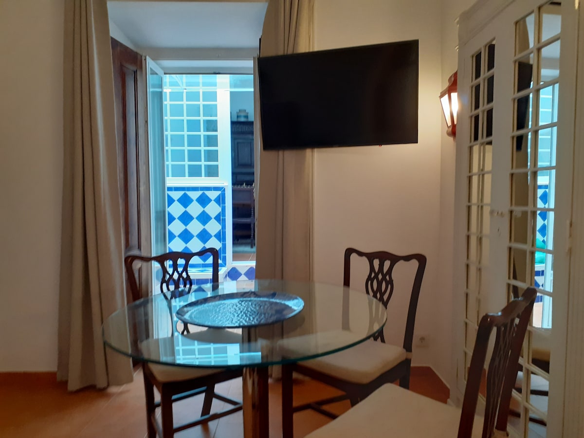 A dining area is presented with a glass table surrounded by four wooden chairs. A large television is mounted on the wall, and natural light enters through the open doors, revealing a glimpse of the shared patio outside.