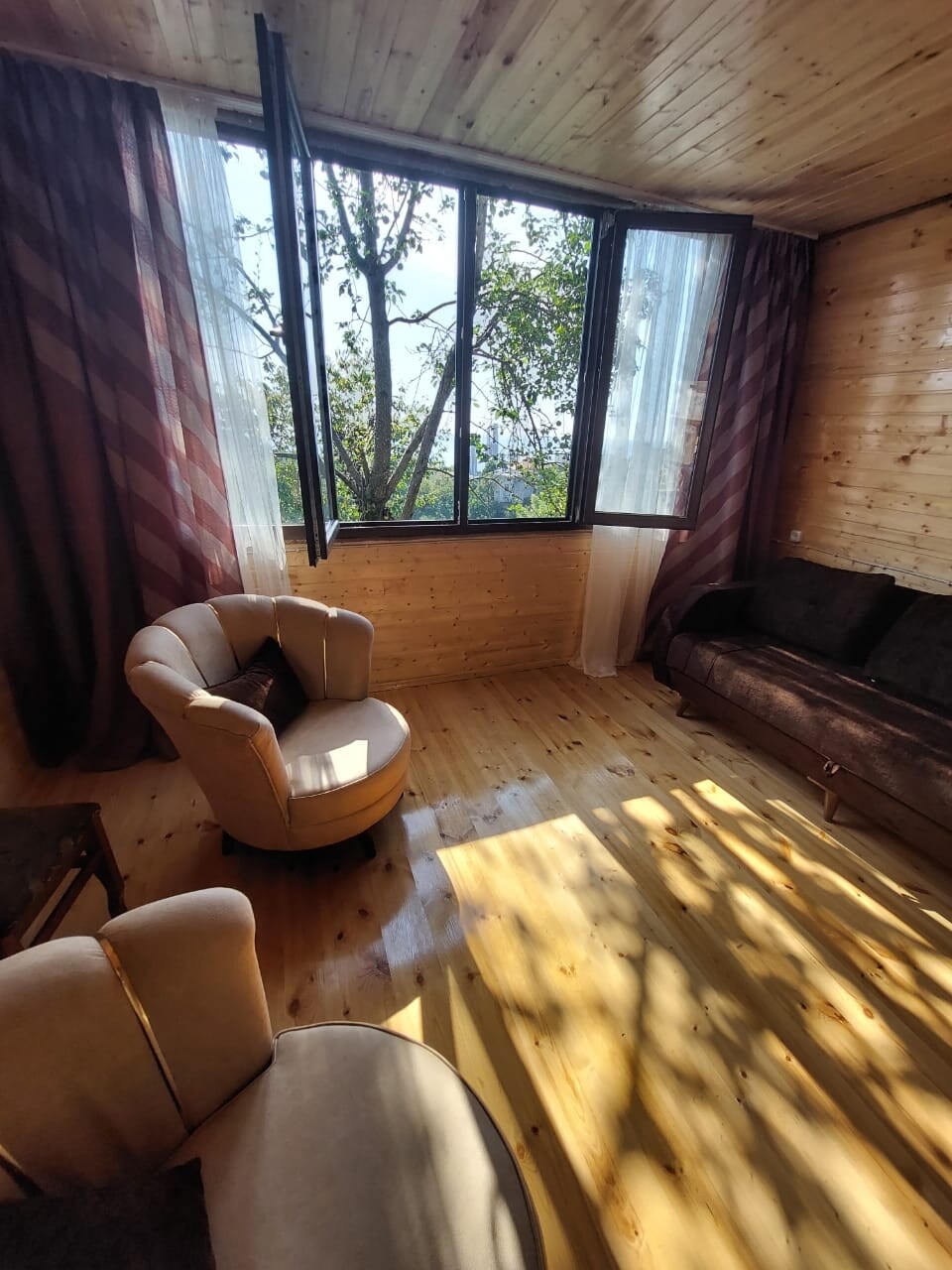 A living area features two seating arrangements, including a cozy armchair and a dark sofa. Large windows allow natural light to fill the space, casting gentle shadows on the wooden floor. Rich curtains frame the windows, contributing to the warm ambiance.