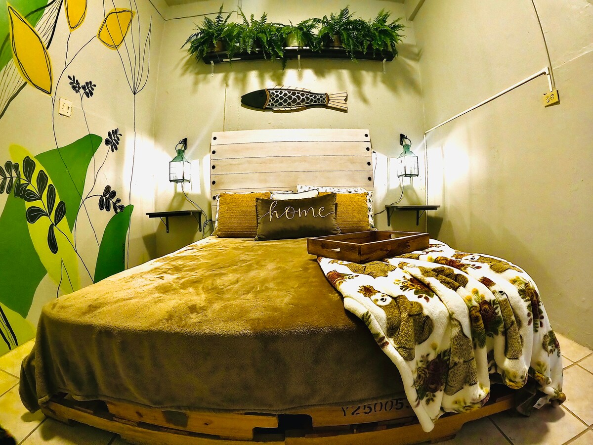 A cozy sleeping area features a spacious bed topped with a soft, neutral blanket and a decorative throw. A wooden headboard adds rustic charm, while wall art of plants and a fish create a nature-inspired ambiance. Gentle lighting is provided by lanterns on either side.