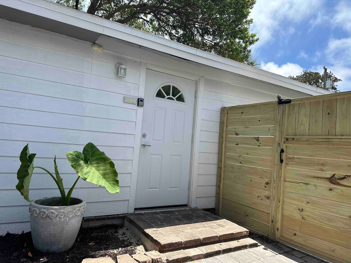 The entrance features a white door with a decorative round window, set against a backdrop of light-colored siding. A large potted plant sits beside the entrance, and a wooden fence borders the area, enhancing the sense of privacy.