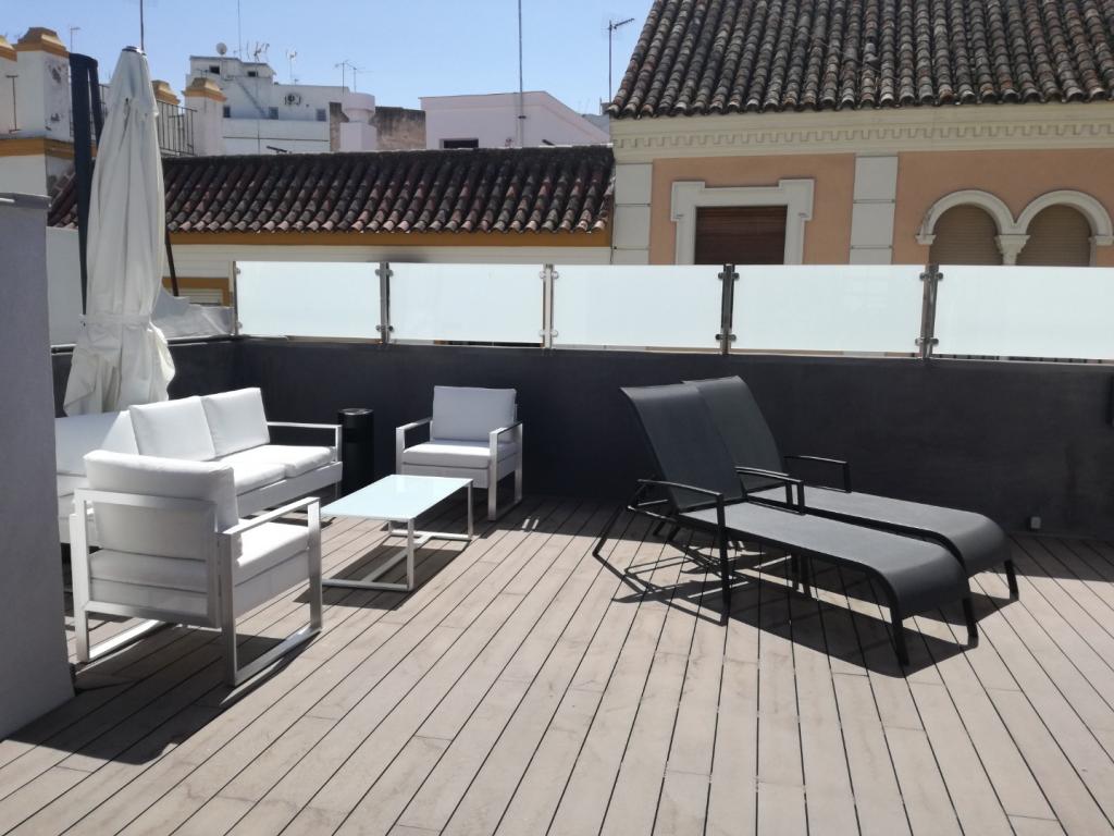 A sunlit terrace features white outdoor seating arranged around a glass-topped table. Two lounge chairs are positioned nearby on a wooden deck, surrounded by a low glass railing. The background shows a glimpse of traditional architectural details.
