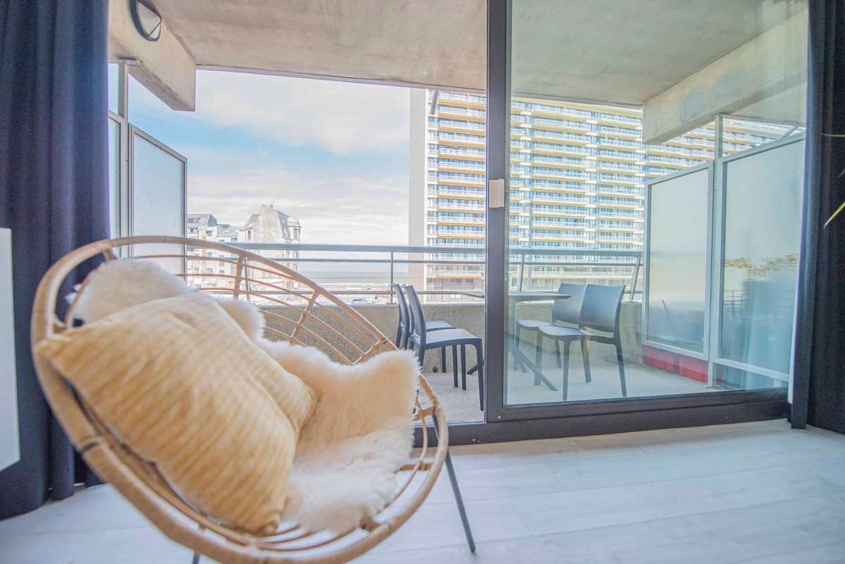 A comfortable chair with soft padding is positioned near a large glass door, providing views of an outdoor balcony with seating. The space is brightened by natural light, with modern buildings visible in the background and clear skies enhancing the serene atmosphere.