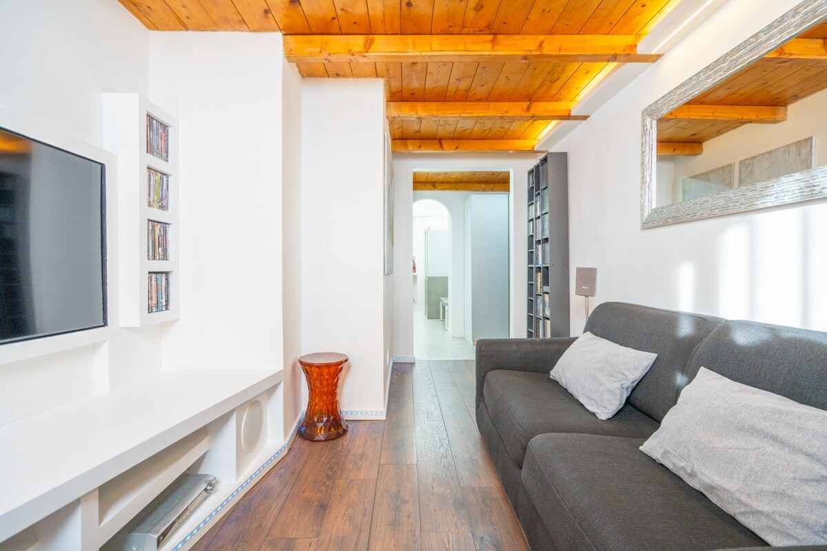 A comfortable living area features a dark grey sofa adorned with light grey pillows. A flat-screen TV rests on a white media unit against the wall. Light wood beams accentuate the ceiling, while a warm-toned side table adds character to the space.