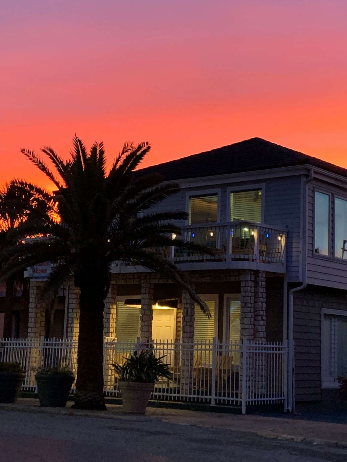 A two-story building with a wraparound balcony is illuminated by the warm glow of sunset. Vibrant hues of orange and pink fill the sky, while a tall palm tree stands in the foreground. Large windows reflect the colorful sky, enhancing the inviting appearance.