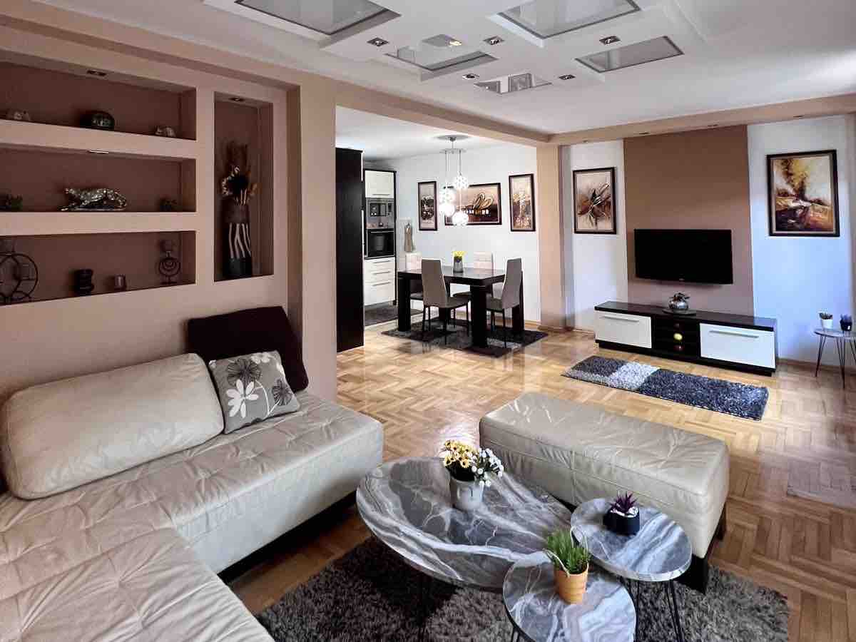 A spacious living area is displayed, featuring a light-colored sectional sofa and unique marble coffee tables. The room is adorned with decorative shelving and framed artworks. A dining area can be seen in the background, accompanied by a modern kitchen and a television unit.