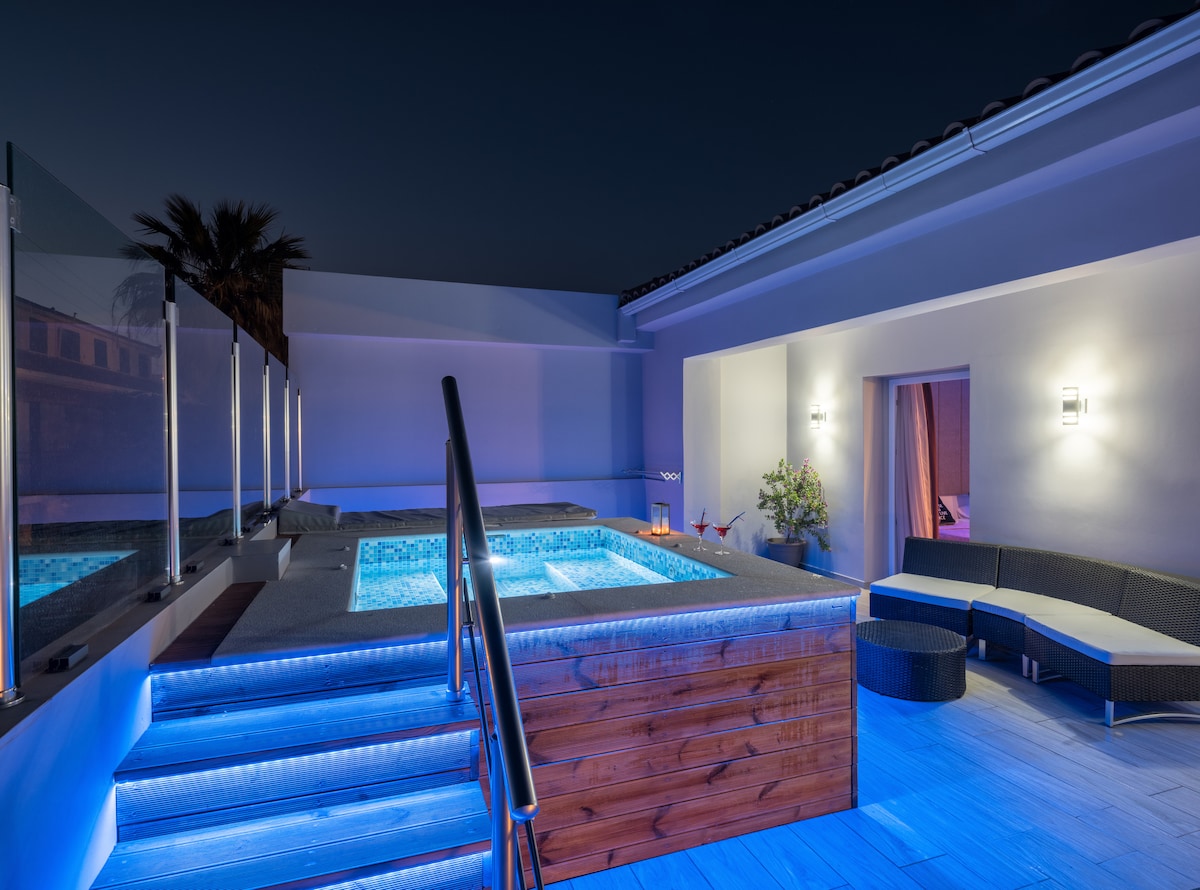 A private balcony area features a Jacuzzi illuminated in blue lighting, surrounded by glass railings. A sectional lounge seating area is positioned nearby, offering a relaxing spot. Soft lighting accentuates the modern design, enhancing the tranquil ambiance at night.