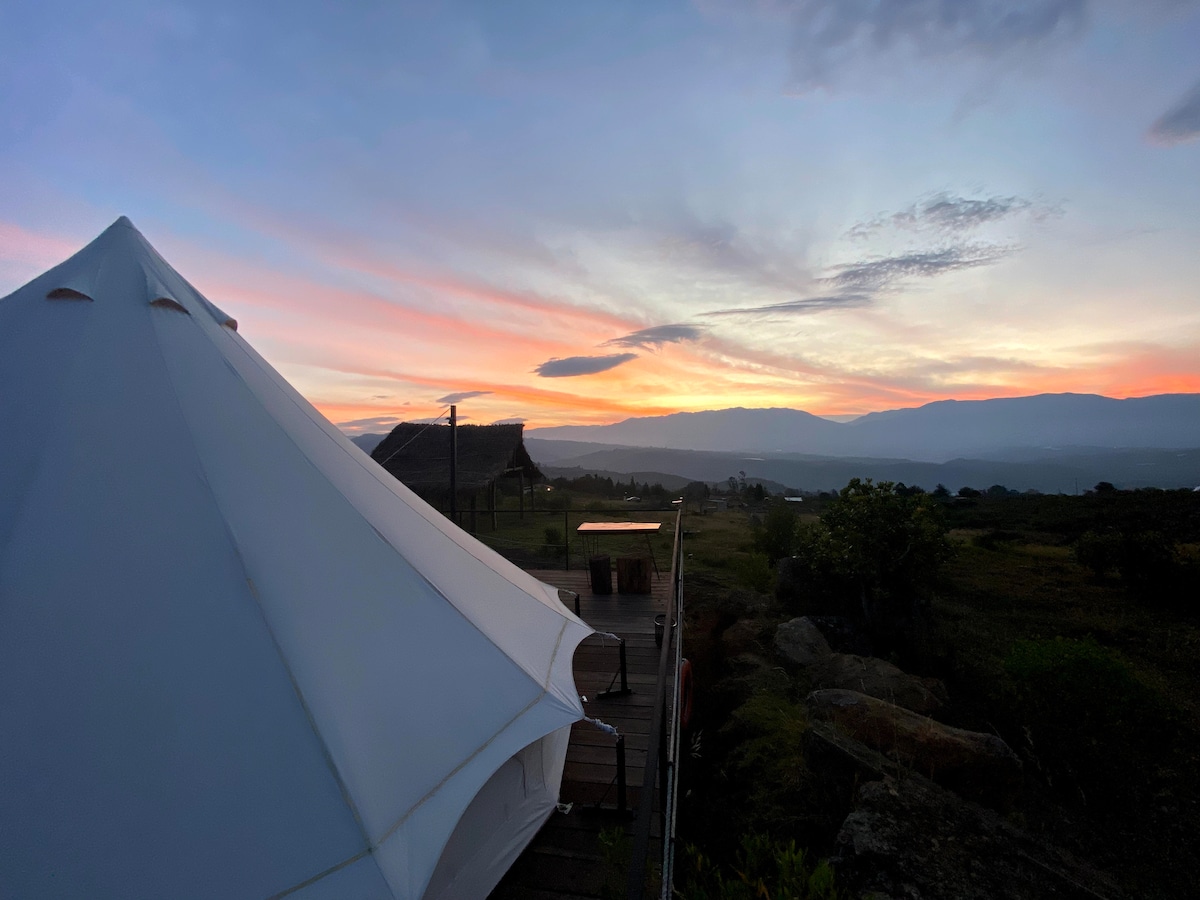 A Tipi-style glamping structure is featured against a vibrant sunset sky. The horizon displays a blend of soft orange and purple hues, while distant mountains provide a serene backdrop. The setting showcases a tranquil natural environment, inviting moments of relaxation and connection with nature.