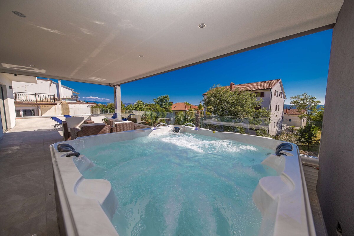 A spacious terrace features a private jacuzzi, inviting relaxation while offering views of the surrounding landscape. Comfortable lounge chairs provide additional seating, and the area is sheltered by a modern roof, ensuring comfort in varying weather conditions.