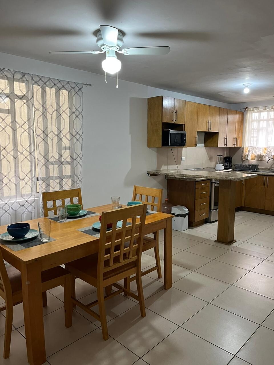 The kitchen and dining area are presented with wooden cabinetry and a small dining table set for four. A ceiling fan provides airflow, while natural light enters through the window covered by sheer curtains, highlighting the tiled flooring and essential kitchen appliances.