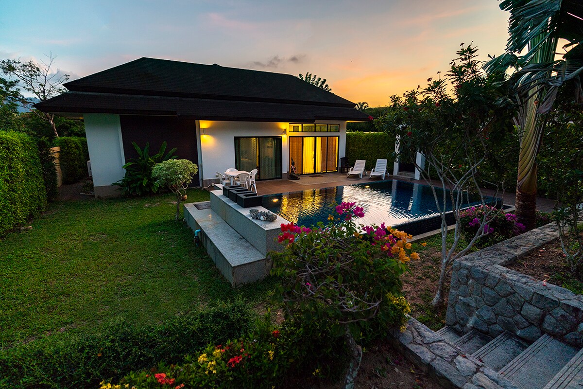 The villa is set within a tranquil garden, featuring a private pool surrounded by vibrant flowers. Sun loungers are positioned beside the pool, with a backdrop of a setting sun. Lush greenery creates a serene atmosphere, enhancing the outdoor space.