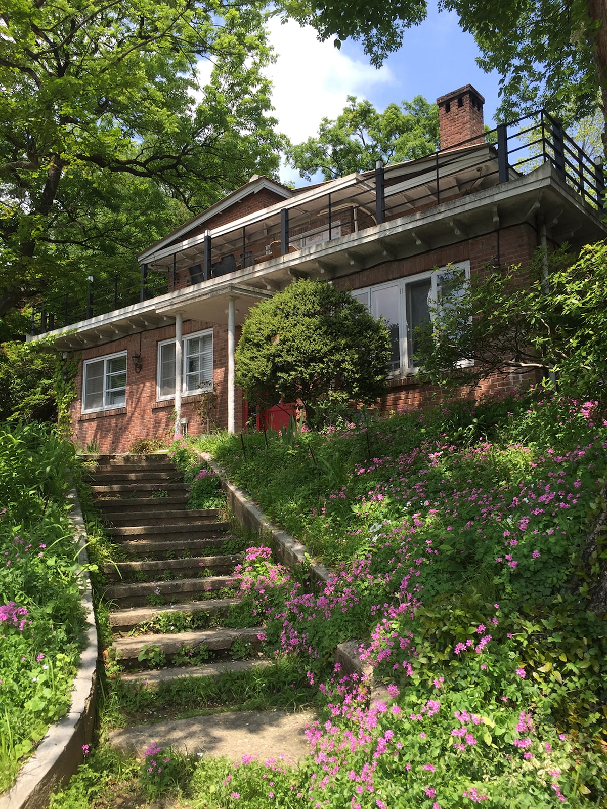 A two-story brick house is set against a backdrop of lush greenery. Stone steps lead to the entrance, flanked by vibrant flowering plants. The upper level features a terrace accessible through large windows, providing a view of the surrounding natural landscape.