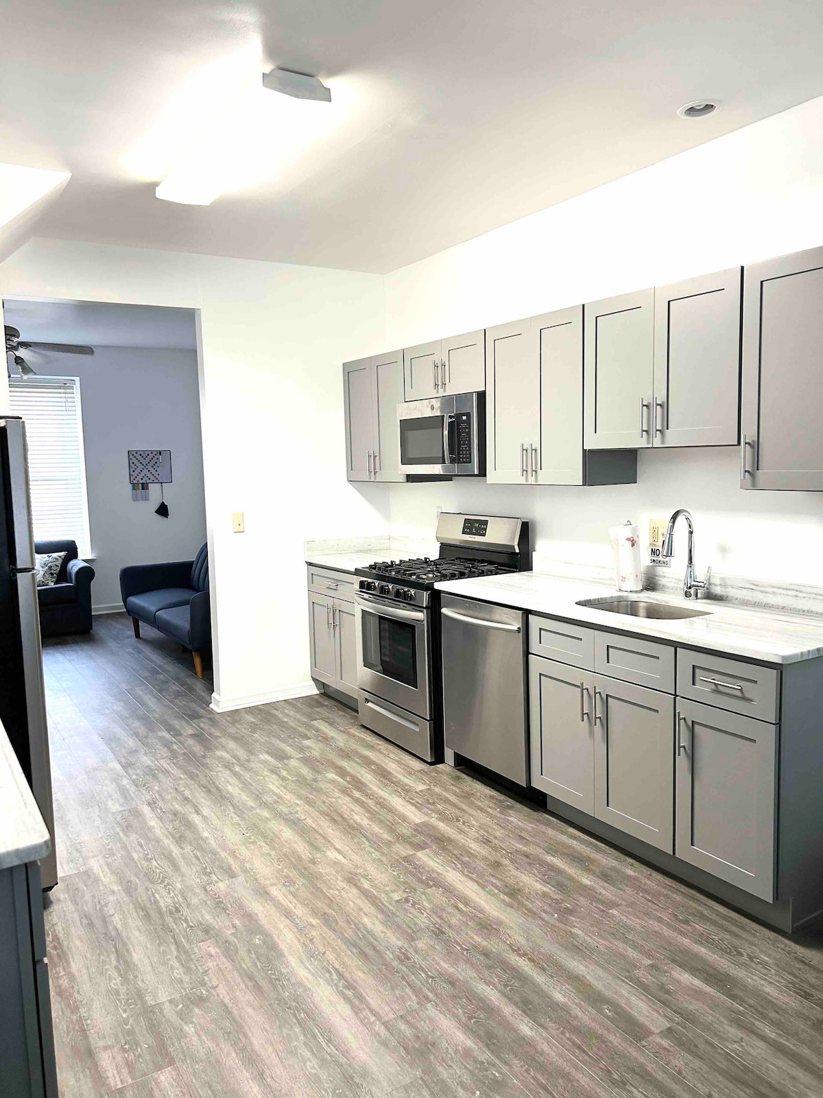 A modern kitchen features grey cabinetry and stainless steel appliances, including a gas range and microwave. A sleek countertop provides ample workspace, with a sink positioned under a window. Light grey flooring completes the contemporary look, with a glimpse of a living area visible through an open doorway.