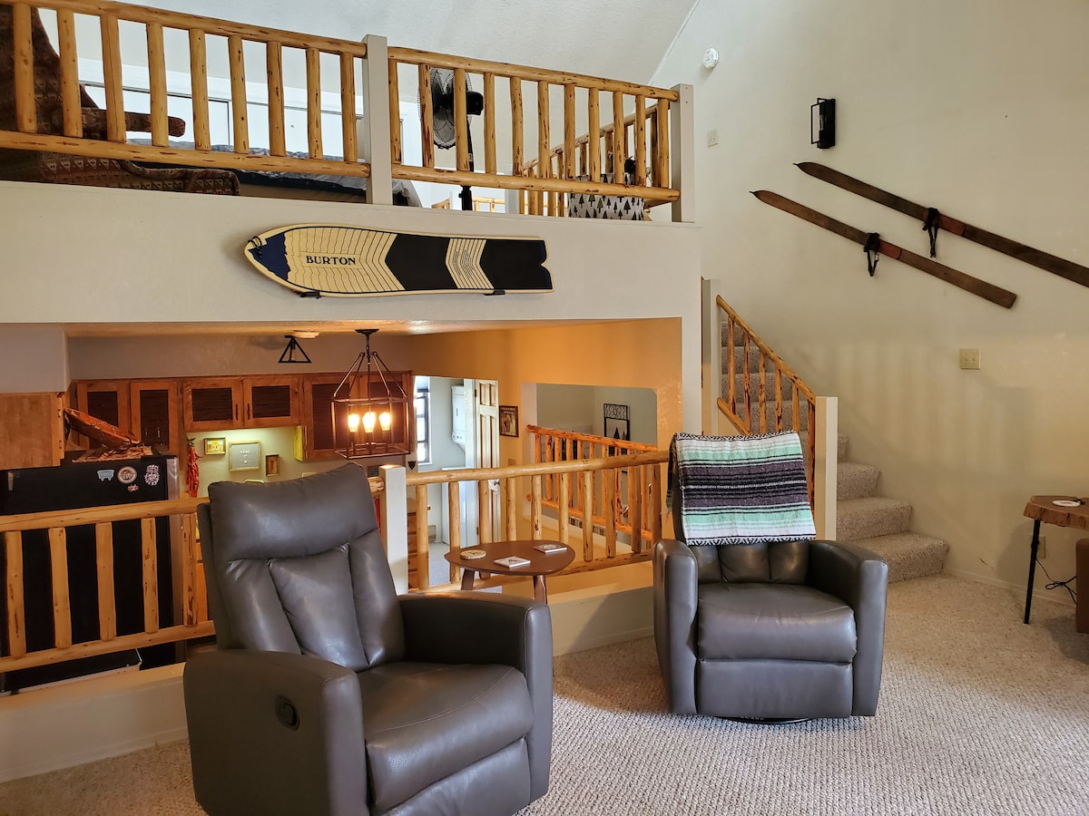 An open living area features a pair of cozy recliners facing a wooden railing that overlooks the loft above. Light gray carpet adds warmth, while rustic ski decor enhances the mountain ambiance. A round wooden table sits adjacent to the reclining chairs, providing additional seating.