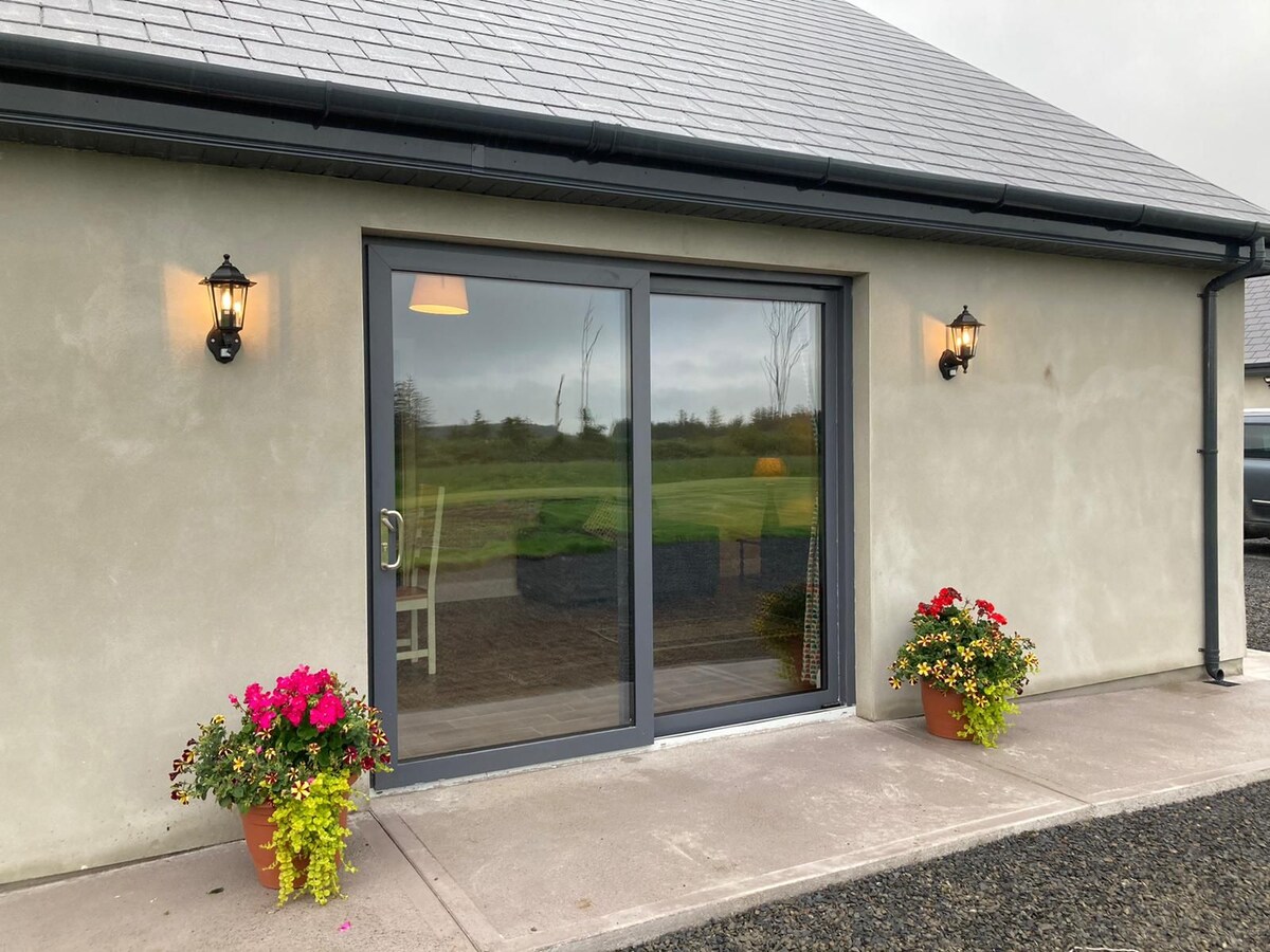 A modern entrance displays large glass doors that invite natural light into the interior. Flanking the entrance, two flower pots filled with colorful blooms add a touch of vibrancy. The surrounding landscape features open countryside views beyond the threshold.