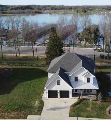 An aerial view of the custom designer home reveals a spacious structure with a grey roof and well-maintained landscaping. The home faces Tim's Ford Lake, with boat docks visible in the background. The surrounding area includes trees and a clear view of the community's green space.