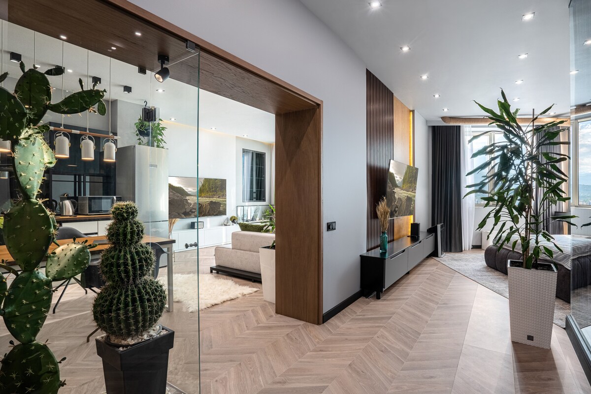 A modern entrance leads to an open living area with large windows allowing natural light to fill the space. Indoor plants are strategically placed, enhancing the fresh ambiance. A dining area with pendant lighting and a living room featuring a wall-mounted television can be seen in the distance.