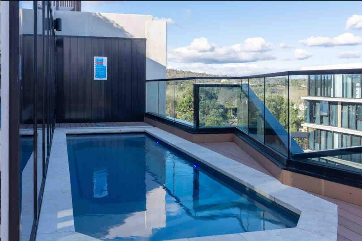 A rooftop swimming pool is framed by glass railings, offering scenic views of the surrounding greenery. The pool features a smooth stone deck and LED lighting, creating a serene atmosphere for relaxation.