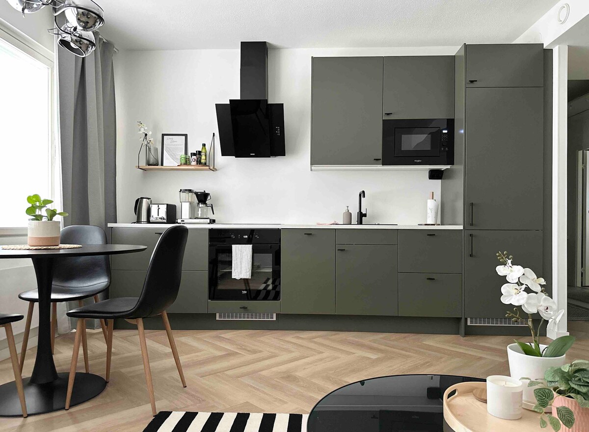 The modern kitchen features sleek green cabinetry, complemented by a black oven and microwave. A round dining table with black chairs is positioned nearby, while natural light filters through the large window, highlighting decorative plants and an elegant orchid on the countertop.