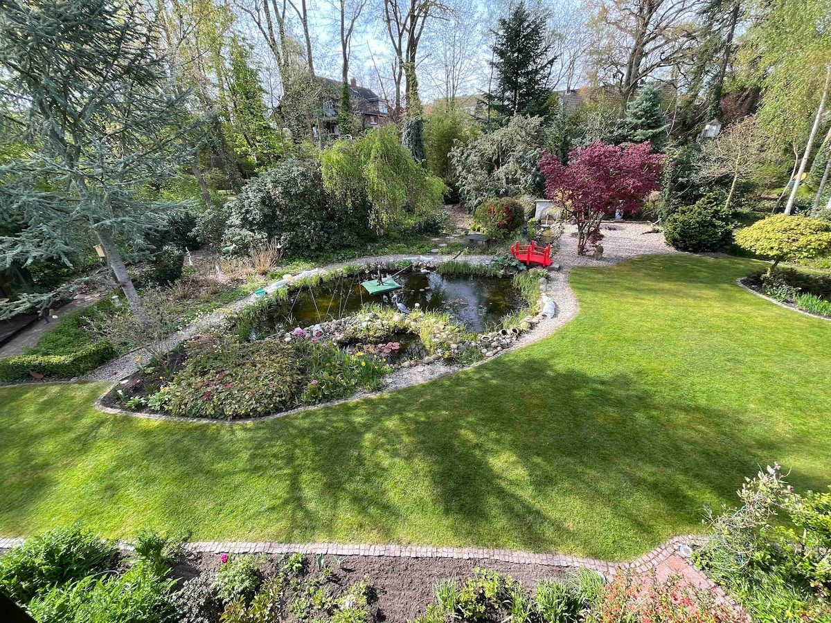 A lush garden is depicted, featuring a winding pathway and a tranquil pond surrounded by diverse greenery and blooming flowers. Two red chairs are positioned near the water, inviting relaxation. Trees and shrubs provide natural shade, enhancing the serene outdoor setting.