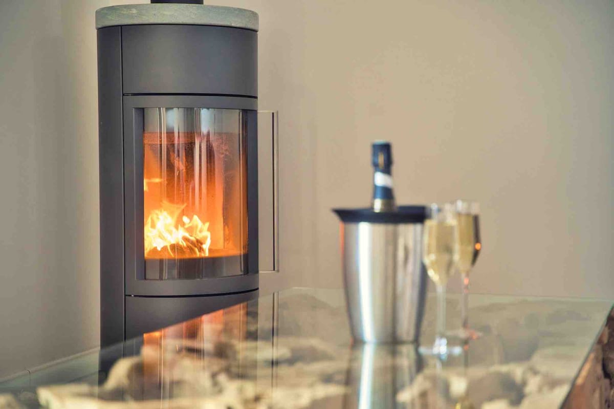 A modern wood stove is showcased, emitting a warm glow from the flames within. A sleek glass table with an elegant marble top is placed nearby, accompanied by a bottle of champagne in an ice bucket and two filled glasses, suggesting a cozy ambiance.