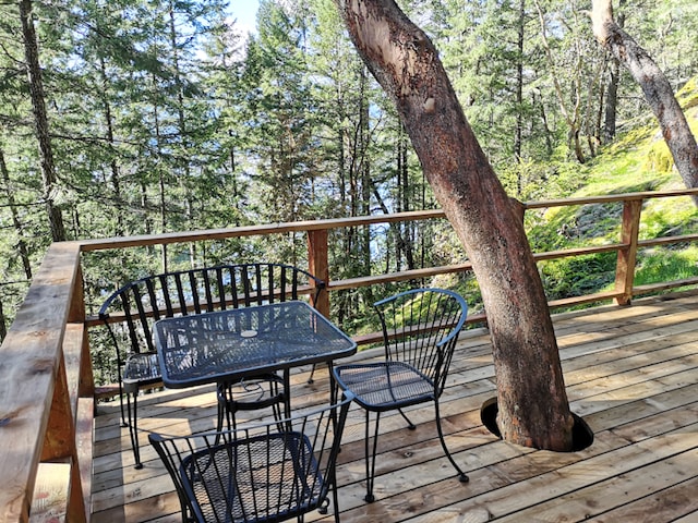 A wooden deck is furnished with a black metal table and four matching chairs. A tree extends through a circular cutout in the deck, surrounded by lush greenery and tall trees. The view beyond highlights the natural beauty of the surrounding forest.