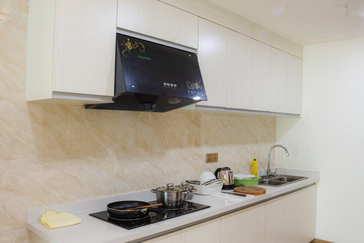 The kitchen features modern cabinetry with a light wood finish and a sleek black range hood above the cooking area. A stovetop is equipped with cookware, while a double sink and countertop provide additional workspace. Common kitchen essentials are visible on the counter.