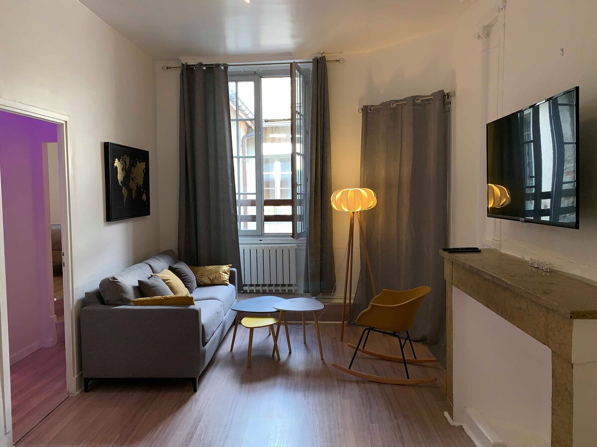 A light-filled living space is presented, featuring a comfortable gray sofa accompanied by yellow accent cushions. A modern coffee table is positioned in front of the sofa, next to a rocking chair. Large windows are framed by long gray curtains, and a flat-screen television is mounted on the wall.