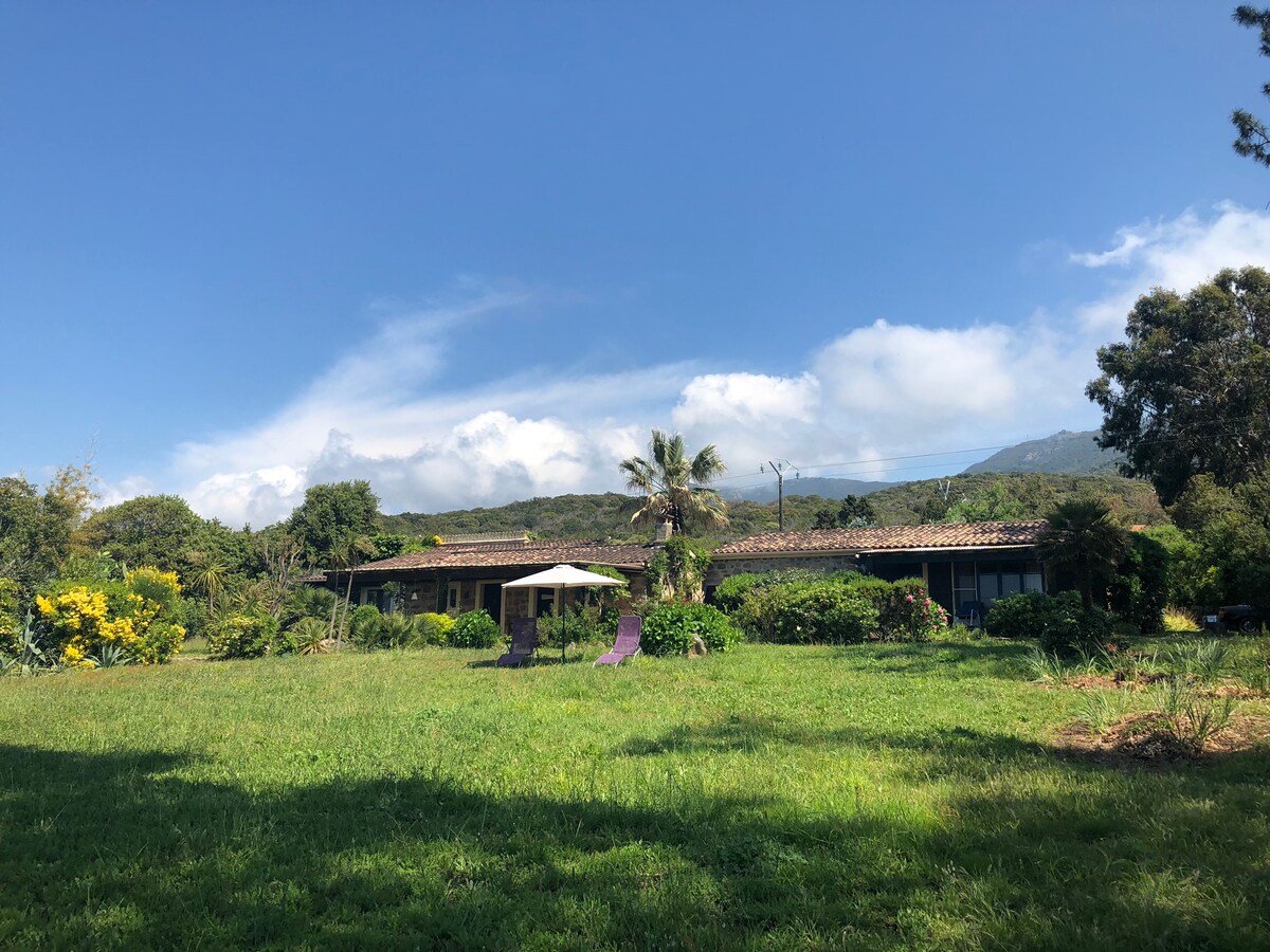 The image showcases a charming villa set within a lush green garden, surrounded by various trees and plants. Sunlight filters through, illuminating the patio area with a small umbrella. The mountains are visible in the background under a partly cloudy sky.