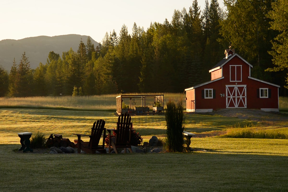 A traditional red barn is set against a backdrop of trees and mountains, with a well-manicured lawn in the foreground. Several Adirondack chairs surround a stone fire pit, inviting relaxation in the serene landscape.