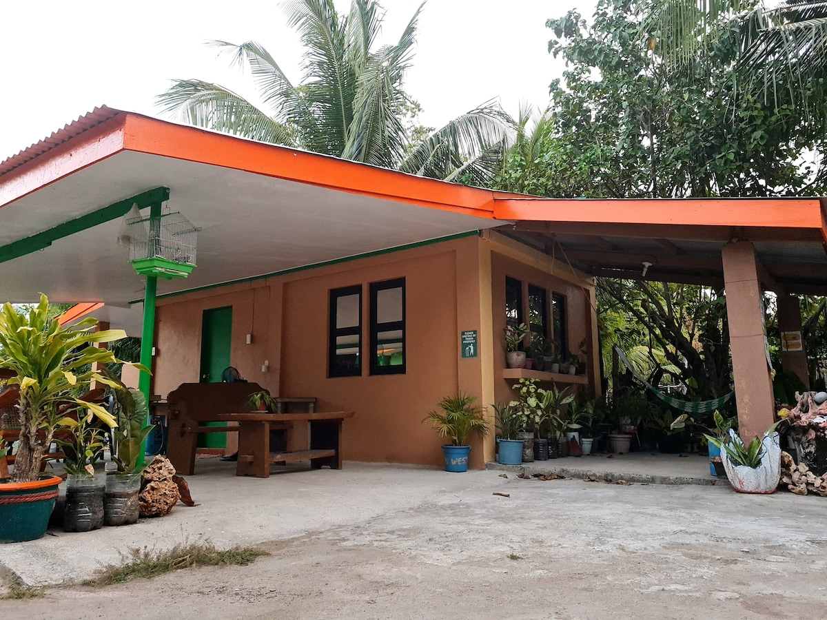 A single-story house features orange overhangs and green trim. Surrounded by lush greenery and potted plants, the outdoor area includes a stone pathway and a picnic table. Large windows allow natural light to brighten the space, offering a welcoming entrance to guests.