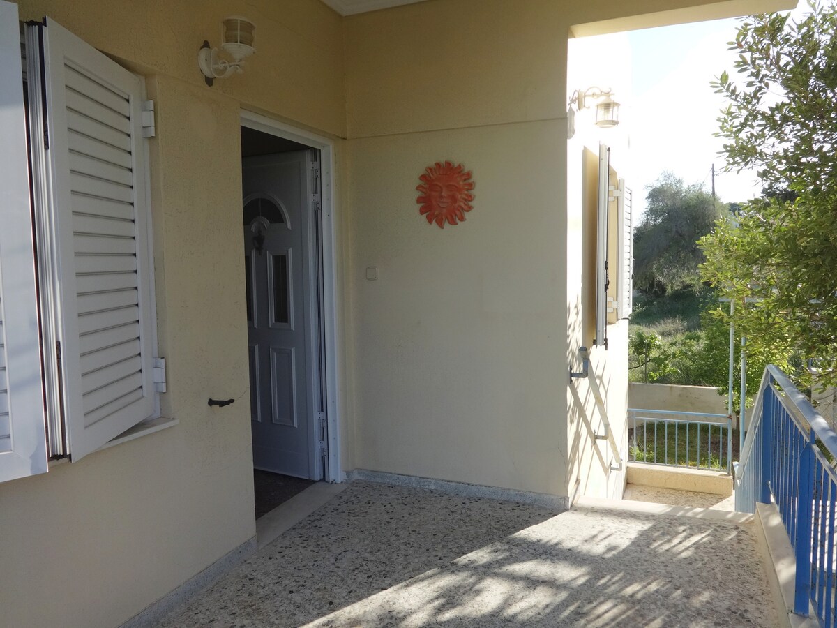 An entrance area is seen, featuring light-colored walls and a smooth stone floor. A decorative wall piece in warm tones adds interest. Bright natural light enters through the open door and windows, creating an inviting space that leads to outdoor views.