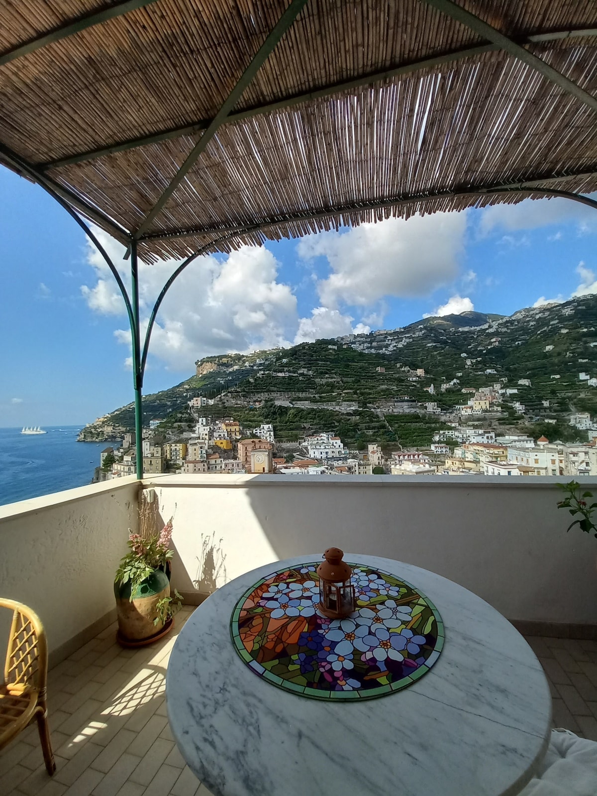 A spacious terrace is adorned with a round marble table featuring a colorful mosaic design. Surrounding greenery adds a natural touch, while the sea view showcases the vibrant town below and the majestic hills in the background. A bamboo canopy provides shade, enhancing the tranquil setting.