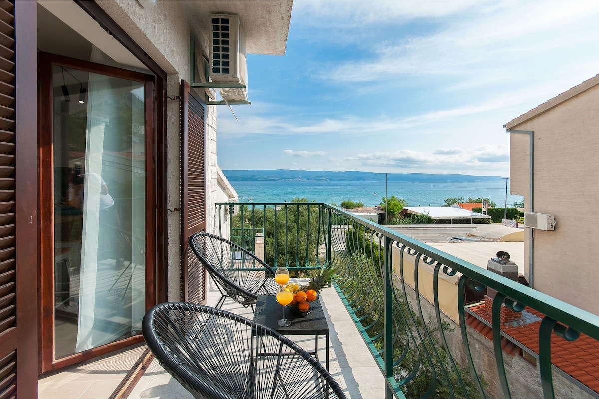 A balcony features two black wicker chairs and a small table adorned with fresh oranges and drinks. The railing offers a clear view of the sea and surrounding landscape, with soft blue skies and distant greenery visible in the background.