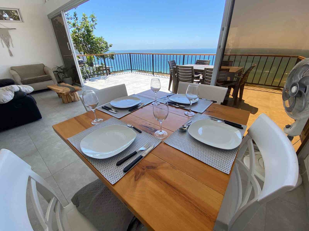 A dining table is set for four, featuring white plates and glassware. Behind it, large glass doors open to reveal a view of the ocean and outdoor seating. Natural light enhances the space, which is complemented by simple, modern decor.