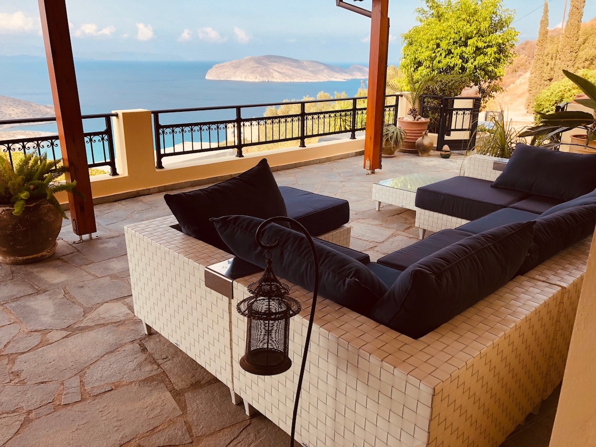 A covered outdoor seating area features modern couches with dark cushions, arranged on a stone-tiled patio. A lantern is positioned on a side table, adding to the relaxed ambiance. The expansive view of the bay is visible in the background, framed by lush greenery.